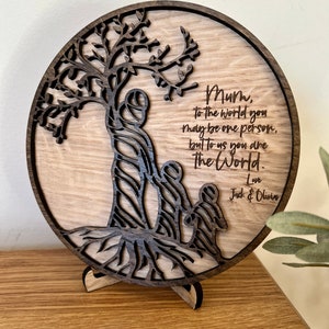 May include: A round wooden plaque with a carved tree design and the text "Mum, to the world you may be one person, but to us you are The World. Love Jack & Olivia".
