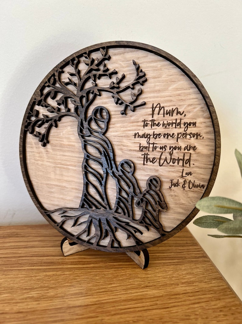 May include: A round wooden plaque with a carved tree design and the text "Mum, to the world you may be one person, but to us you are The World. Love Jack & Olivia".