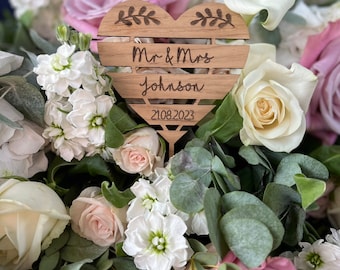 May include: A wooden heart-shaped sign with the text "Mr. & Mrs. Johnson 21.08.2023" engraved on it. The sign is surrounded by a bouquet of white, pink, and cream flowers.