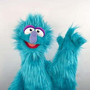 May include: A bright turquoise fuzzy hand puppet with long synthetic fur, large white eyes with black pupils, purple eyelids, a purple nose, and an open mouth showing a red interior. The puppet has one arm raised in a waving gesture.