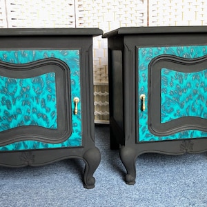 May include: Two black bedside tables with teal and gold patterned doors and gold handles. The bedside tables have ornate legs and are sitting on a blue carpet.