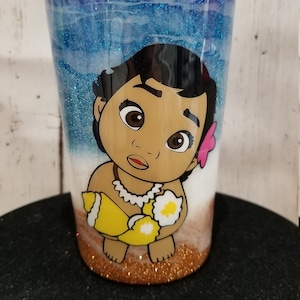 May include: A clear tumbler with a blue and white glitter design featuring a cartoon image of a baby Moana holding a seashell. The bottom of the tumbler is filled with gold glitter.
