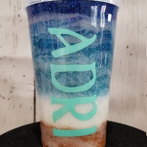 May include: A clear plastic tumbler with a blue, white, and gold glitter design. The tumbler has a turquoise name "Adri" on the side.