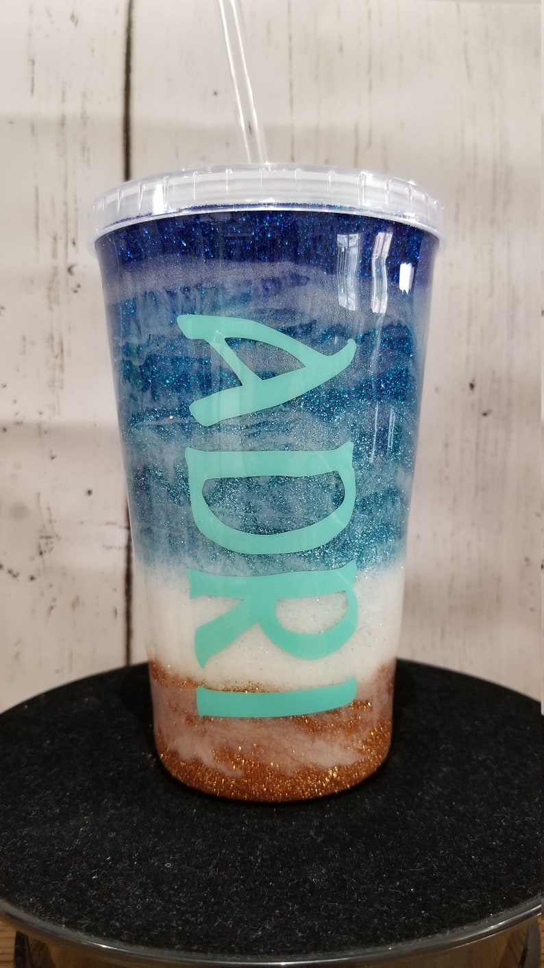 May include: A clear plastic tumbler with a blue, white, and gold glitter design. The tumbler has a turquoise name "Adri" on the side.