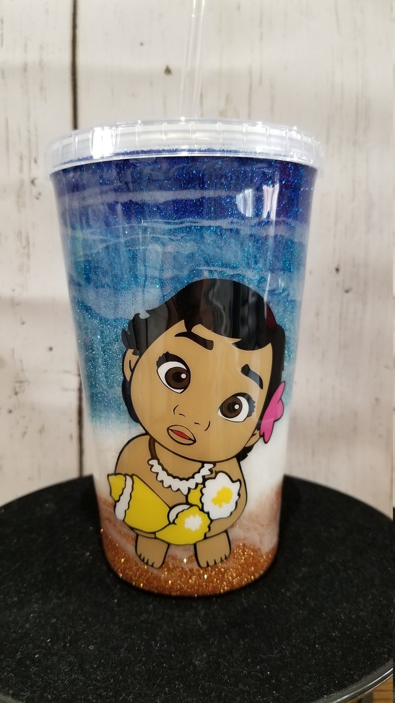 May include: A clear tumbler with a blue and white glitter design featuring a cartoon image of a baby Moana holding a seashell. The bottom of the tumbler is filled with gold glitter.