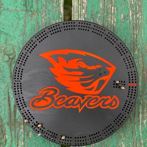 May include: A round, black cribbage board featuring the Oregon State Beavers logo in orange. The board has numerous holes for pegs and the word "Beavers" in orange script. The board is on a weathered green wooden surface.