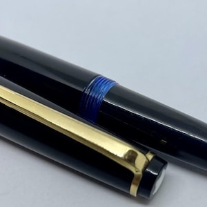 May include: A black fountain pen with a gold-colored nib and accents. The pen's cap is off, revealing the nib and the pen's body. A blue band is visible near the top of the pen. The pen is a writing instrument.