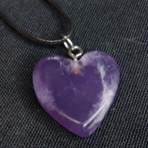 May include: A heart-shaped pendant necklace featuring a polished purple stone with white marbling. The pendant is suspended from a black cord necklace with a silver-colored clasp. The necklace is displayed against a dark background.