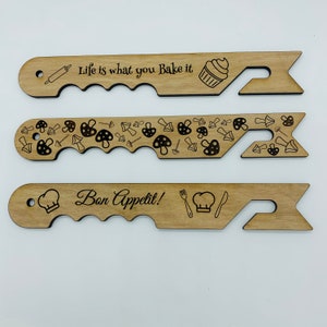 May include: Three wooden kitchen utensil holders with different designs. The top holder has a rolling pin, cupcake, and the text "Life is what you bake it". The middle holder has a mushroom pattern. The bottom holder has a chef's hat, fork, knife, and the text "Bon Appetit!"