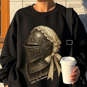 May include: Black sweatshirt with a detailed illustration of a medieval helmet with a lace-trimmed hood and bow. The text "FIRST, YOU UNRAVEL" is printed below the helmet. The sweatshirt is paired with black shorts.