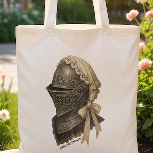 May include: Cream-colored tote bag featuring a detailed illustration of a medieval knight's helmet with a lace veil and bow. The text "FIRST, YOU UNRAVEL" is printed at the bottom. The bag has two handles and is set against a garden backdrop.