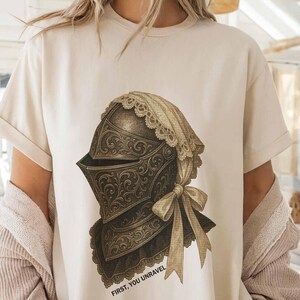 May include: Cream-colored t-shirt featuring a detailed illustration of a medieval knight's helmet with ornate scrollwork and a lace-trimmed veil. The text "FIRST, YOU UNRAVEL" is printed below the helmet.