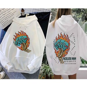 Puede incluir: Sudadera con capucha blanca con un gr&aacute;fico de tres calaveras turquesas con huesos cruzados y un coraz&oacute;n, rodeadas de llamas y sostenidas por una mano esquel&eacute;tica. La parte trasera tiene el texto "FACELESS MAN SECURITY".