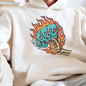 Puede incluir: Sudadera con capucha color crema con un gr&aacute;fico de tres calaveras azules en llamas, sostenidas por una mano esquel&eacute;tica. Las calaveras tienen detalles de huesos cruzados y corazones. El dise&ntilde;o est&aacute; centrado en la parte delantera de la sudadera.
