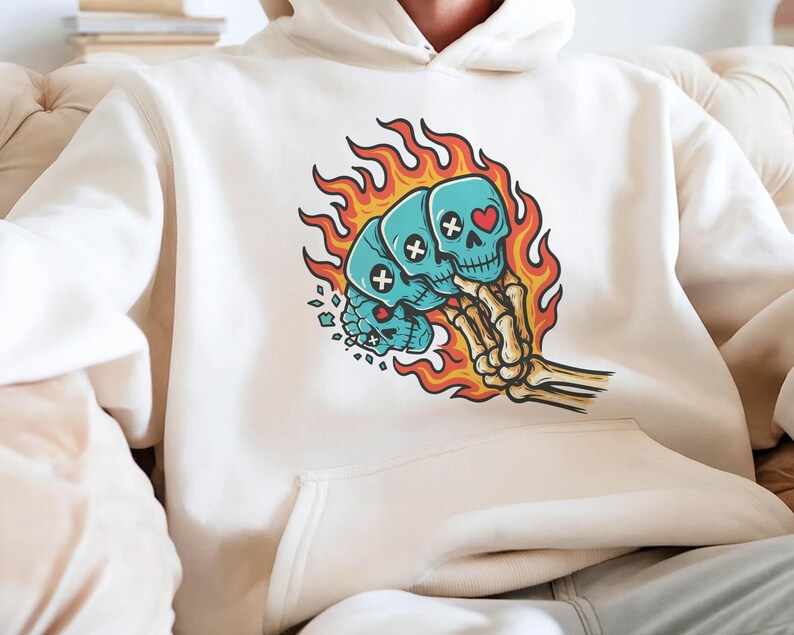 Puede incluir: Sudadera con capucha color crema con un gr&aacute;fico de tres calaveras azules en llamas, sostenidas por una mano esquel&eacute;tica. Las calaveras tienen detalles de huesos cruzados y corazones. El dise&ntilde;o est&aacute; centrado en la parte delantera de la sudadera.