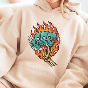 Puede incluir: Sudadera con capucha beige con un gr&aacute;fico de tres calaveras turquesas en llamas, sostenidas por una mano esquel&eacute;tica. Las calaveras tienen ojos de huesos cruzados o en forma de coraz&oacute;n. Las llamas son naranjas y amarillas.
