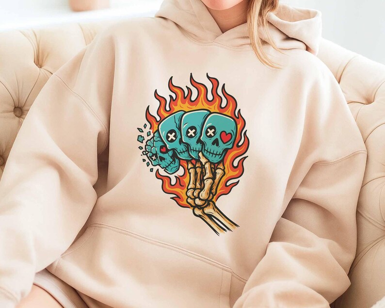 Puede incluir: Sudadera con capucha beige con un gr&aacute;fico de tres calaveras turquesas en llamas, sostenidas por una mano esquel&eacute;tica. Las calaveras tienen ojos de huesos cruzados o en forma de coraz&oacute;n. Las llamas son naranjas y amarillas.