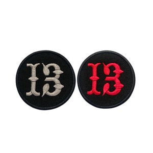 Könnte beinhalten: Zwei schwarze gestickte Patches mit der Zahl 13 in einer stilisierten Schriftart. Ein Patch hat silbernen Faden und der andere roten Faden.