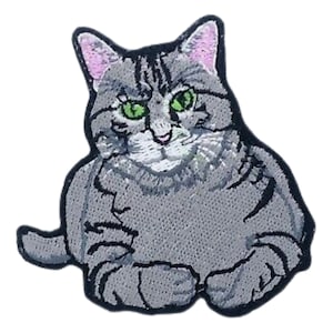 May include: A gray and white tabby cat with green eyes is sitting with its paws tucked under its body. The cat is looking directly at the viewer. The cat is embroidered on a fabric patch.