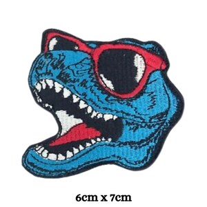 May include: Blue dinosaur head with red sunglasses and white teeth. The dinosaur is embroidered on a black fabric patch. The patch measures 6 cm by 7 cm.