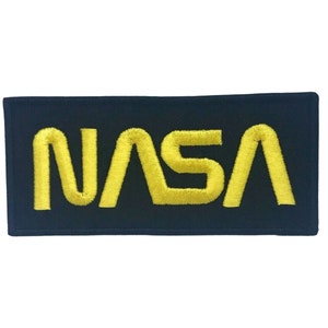 Puede incluir: Un parche bordado negro con el logotipo de la NASA en amarillo.