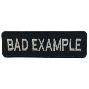 May include: A rectangular black patch with the words "BAD EXAMPLE" embroidered in white block letters. The patch has a simple, clean design, suitable for application on clothing or accessories.
