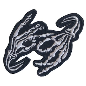 Op de afbeelding: Zwart-witte geborduurde patch van twee skeletachtige handen die naar elkaar reiken.