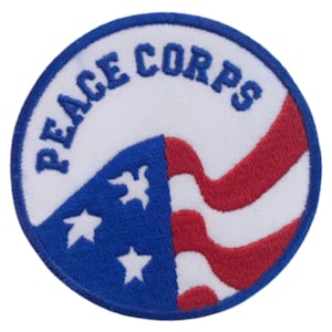May include: A round, white patch with the words "Peace Corps" in blue lettering around the edge. The center of the patch features a stylized American flag with red and white stripes and blue stars.