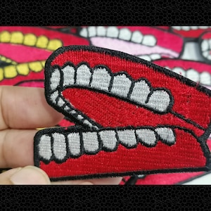 May include: Red embroidered patch of a set of teeth, with white teeth and black outlines. The teeth are in a mouth shape, with the top teeth showing and the bottom teeth showing.