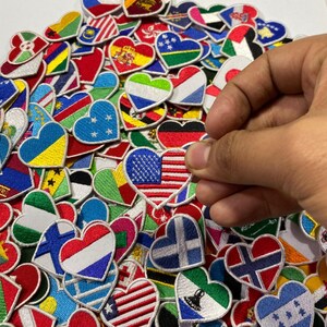Puede incluir: Una colección de parches bordados en forma de corazón con banderas de varios países. Los parches están dispuestos en un patrón aleatorio y son principalmente rojos, blancos y azules. Una mano sostiene un parche con la bandera estadounidense.
