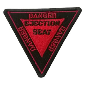 Puede incluir: Un parche triangular negro con un triángulo rojo en el interior. El triángulo rojo tiene las palabras "DANGER EJECTION SEAT DANGER" escritas alrededor en negro.