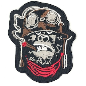Peut inclure: Patch brodé représentant un singe de dessin animé portant un casque d'aviateur en cuir marron avec des lunettes, un bandana rouge et fumant un cigare. Le singe a une expression sérieuse et est sur un fond noir.