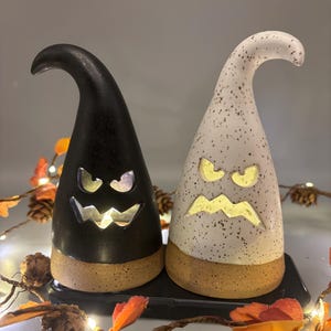 May include: Two ceramic Halloween decorations shaped like witch hats. One is black, the other is white with black speckles. Both have carved-out faces that glow with yellow light. They sit on a black tray with a brown base.