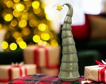 May include: A decorative, green ceramic Christmas tree with a star on top. The tree is on a red and black plaid tablecloth, with wrapped gifts and a lit Christmas tree in the background. The tree is approximately 10 inches tall.