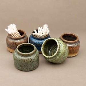 May include: A set of five small, round ceramic pots with speckled glazes in shades of brown, blue, and green. Two of the pots are filled with white cotton swabs. The pots have a smooth, rounded shape with a narrow opening at the top.