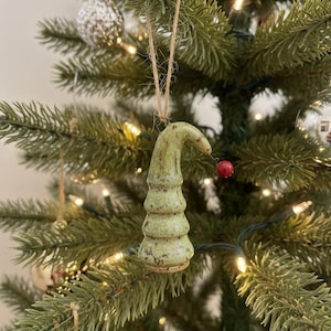 May include: A green ceramic Christmas tree ornament with a curved top and a small red berry. The ornament is suspended by a natural twine string and hangs on a green Christmas tree.