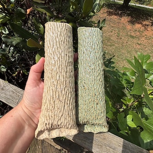May include: Two cylindrical ceramic vases with a textured, bark-like surface. One vase is a light cream color with subtle brown speckles, while the other is a muted sage green. Both pieces feature a vertical, rugged pattern resembling tree bark.