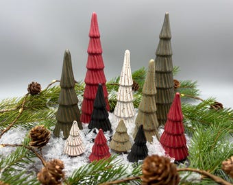 May include: A collection of ceramic Christmas trees in various sizes and colours, including red, green, white, and black. The trees are arranged on a bed of faux snow and surrounded by pine branches.