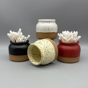 May include: Four small ceramic jars in black, white, cream, and red, each with a speckled design and a brown base. Two jars hold cotton swabs. The jars are arranged on a gray surface.