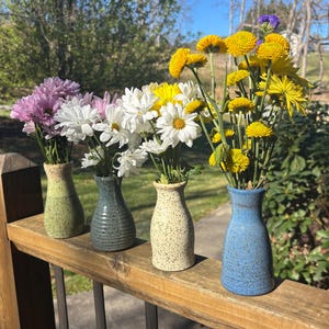 May include: Four small ceramic vases in shades of green, blue, and cream, each holding a different arrangement of fresh flowers. The flowers include purple, white, and yellow blooms. The vases are displayed on a wooden railing.