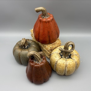 May include: Four ceramic pumpkins in shades of orange, brown, green, and cream, each with a textured surface and a curved stem. One pumpkin rests on a small bale of hay. The pumpkins are arranged on a gray background, evoking a fall harvest theme.