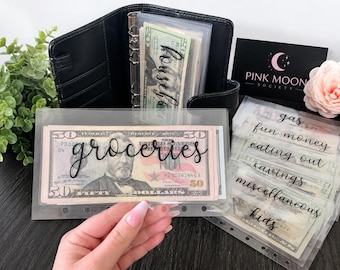 May include: Black leather binder with clear plastic budget envelopes labeled "groceries" and "household." Additional envelopes are labeled "gas," "fun money," "eating out," "savings," "miscellaneous," and "kids." A Pink Moon Society business card is also visible.