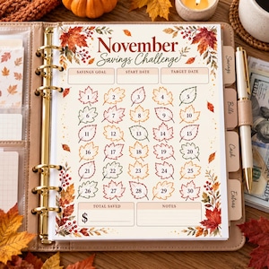May include: A November Savings Challenge tracker sheet inside a gold-ringed binder. The page features autumn leaf illustrations, a grid of thirty numbered leaves for tracking, and fields for savings goal, dates, total saved, and notes in dark red text.