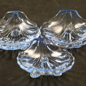 May include: Three light blue, seashell-shaped glass dishes. Each dish has a scalloped edge and a detailed, ridged interior. The dishes are arranged on a dark surface, showcasing their delicate design and color.