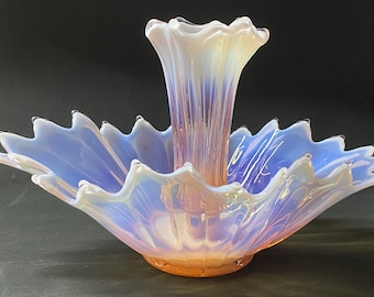 May include: An opalescent glass centerpiece bowl featuring a central vertical vase component. The piece has a ruffled, scalloped edge and displays a translucent blue and white finish that transitions into a warm peach hue at the base.