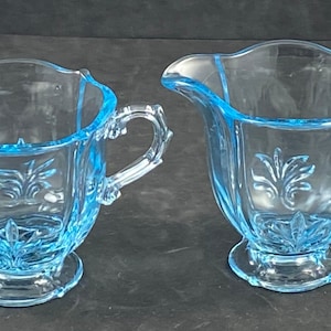 May include: A light blue glass creamer and sugar bowl set. The sugar bowl has two handles, and both pieces feature a floral design. The set is made of clear glass with a light blue tint.