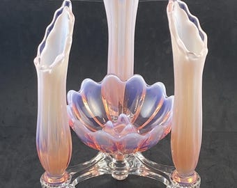 Pode incluir: Um centro de mesa de vidro opalescente de três peças com uma taça central de borda recortada e três vasos altos em forma de trombeta. O vidro apresenta um acabamento iridescente rosa e azul, apoiado numa base de vidro moldado transparente.