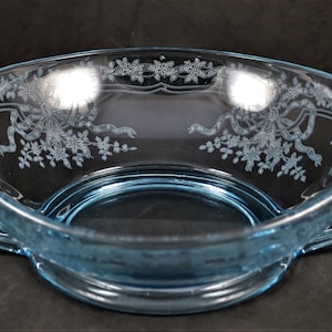 May include: A light blue glass bowl with etched floral and ribbon designs. The bowl has two curved handles with spiral ends. The bowl is likely vintage and is suitable for serving or display.