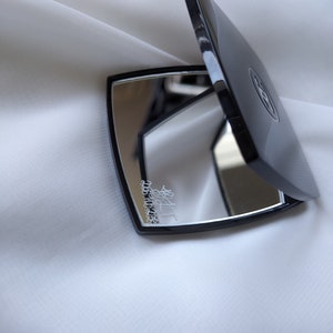 May include: A black, open compact mirror with a reflective surface. The mirror has a square shape with rounded edges and a black frame. The compact is resting on a white, textured surface. The compact has a logo on the top.