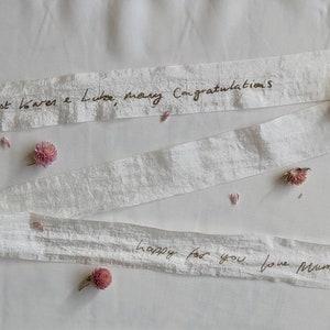 May include: White ribbon with handwritten message "Dearest Laura & Luke, many congratulations happy for you love Mum x" on a white background with pink flowers.